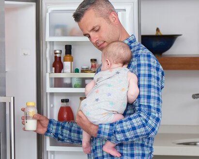 Un père tient son bébé dans la cuisine pendant qu'il sort un biberon du réfrigérateur pour le réchauffer.